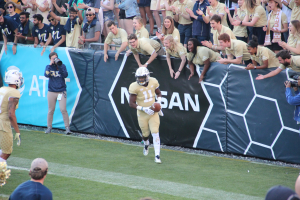 Ein Fußballspieler läuft mit dem Ball auf einem grünen Rasen, mit einer Menge Menschen im Hintergrund, einige halten Kameras und tragen Helme, und ein Banner mit Text im Hintergrund.