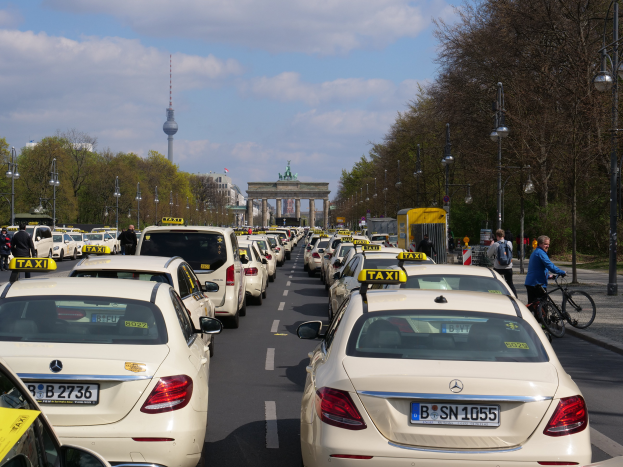 Eine lange Reihe von Taxis, die auf der Seite einer belebten Straße in Berlin, Deutschland, geparkt sind, mit Fahrzeugen, Radfahrern und Fußgängern, flankiert von Laternenmasten, Bäumen und Gebäuden, einschließlich eines Bogens und eines Turms.
