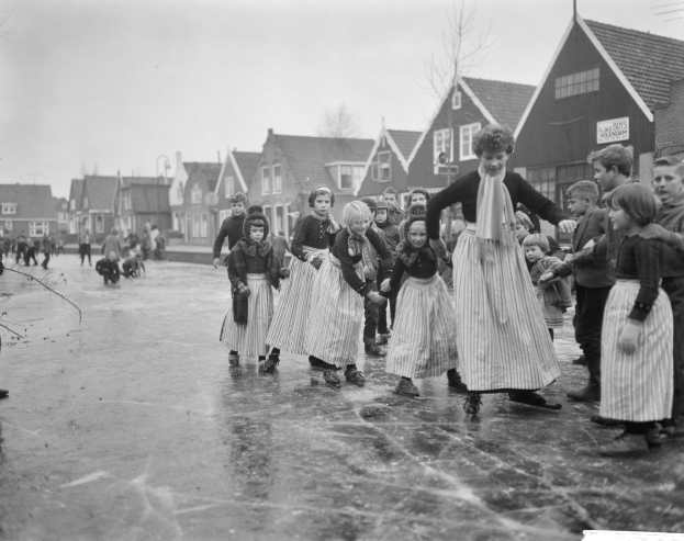 Eine Gruppe von Kindern, die auf einer Eisbahn vor einem Gebäude mit Fenstern, Bäumen und einem bewölkten Himmel Schlittschuh laufen, dargestellt in Schwarz-Weiß.