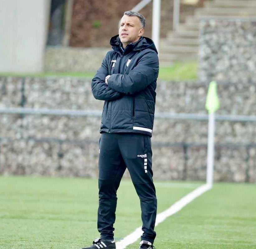 Ein Mann in schwarzer Jacke und Hose steht auf einem Fußballfeld mit verschränkten Armen, der wie ein Trainer während eines Trainings erscheint, mit einem Zaun, Treppen und einer Wand im Hintergrund.