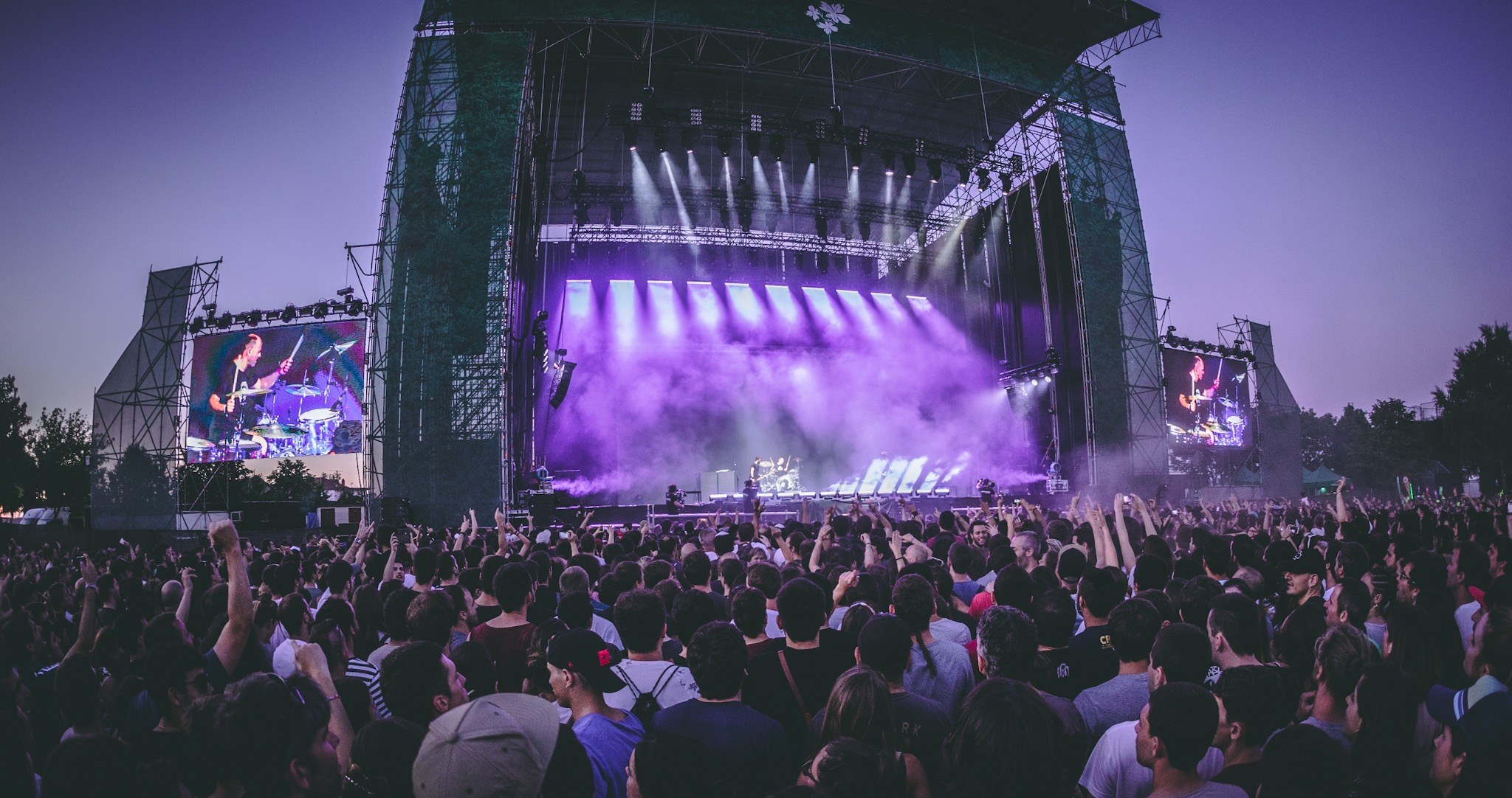 Eine große Menschenmenge auf einem Musikfestival schaut auf der hell erleuchteten Bühne mit Instrumenten und Bildschirmen, die vor einem Gebäude, Bäumen und einem klaren blauen Himmel aufgebaut sind.