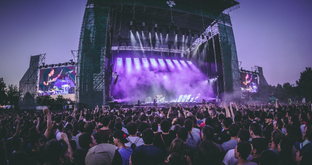 Eine große Menschenmenge auf einem Musikfestival schaut auf der hell erleuchteten Bühne mit Instrumenten und Bildschirmen, die vor einem Gebäude, Bäumen und einem klaren blauen Himmel aufgebaut sind.