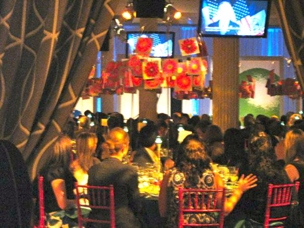 Eine Gruppe von Menschen, die an Tischen mit Essen, Fernsehern, Lichtern, hängenden Papieren und Vorhängen sitzt, in einer Art Party-Atmosphäre.