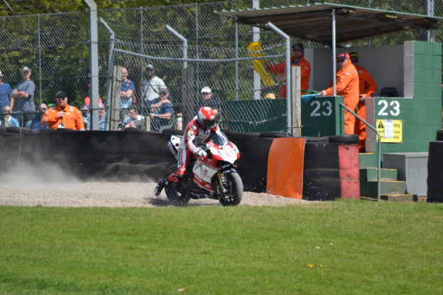 Eine Person in Helm und Handschuhen fährt ein Motorrad über ein grasbewachsenes Feld, mit Zuschauern in Kappen und Schutzbrillen, einem Zaun, Treppen mit Geländern und einem nummerierten Schuppen im Hintergrund.