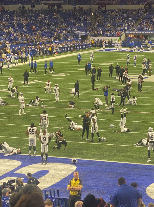 Ein Fußballspiel wird in einem Stadion gespielt, während Menschen auf dem Boden stehen und einige auf Stühlen im Hintergrund sitzen und eine Person eine Kamera hält, was auf eine Live-Übertragung hinweist.