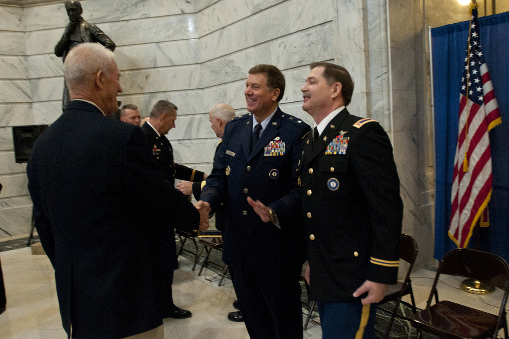 Menschen schütteln sich die Hände in einem Raum mit Stühlen, einer Statue neben einer Wand, einer Flagge und einem Vorhang auf der rechten Seite.