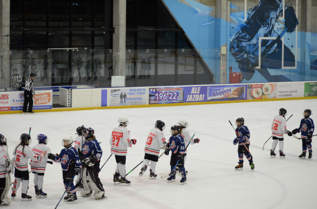 Gruppe junger Menschen, die Eis Hockey auf einer Indoor-Eisbahn spielen, mit Helmen und Stöcken, sowie einer Person auf der linken Seite und Bannern, Säulen und Glaswänden im Hintergrund.