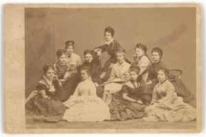 Eine Gruppe von Frauen, die auf dem Boden sitzen und eine Frau in der Mitte steht, die direkt in die Kamera schaut.