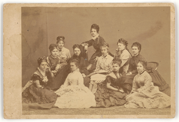 Eine Gruppe von Frauen, die auf dem Boden sitzen und eine Frau in der Mitte steht, die direkt in die Kamera schaut.