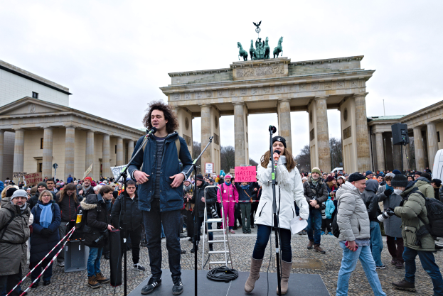 Eine Gruppe von Menschen auf einer Bühne vor dem Brandenburger Tor in Berlin, Deutschland, mit zwei Personen, die Mikrofone halten, und einer weiteren Person mit einer Kamera, sowie einer Leiter im Hintergrund und Gebäuden, Bäumen und einem klaren blauen Himmel.
