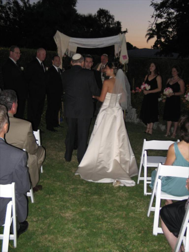 Eine Gruppe von Menschen steht bei einer Hochzeit, wobei Frauen auf der rechten Seite Blumensträuße halten, während andere auf Stühlen sitzen und Bäume im Hintergrund zu sehen sind.