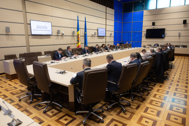 Große Gruppe von Menschen an einem langen Tisch mit Mikrofonen und Papieren in einer formellen Sitzung mit Flaggen der Europäischen Union und Deckenleuchten.