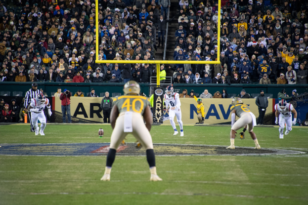 Fußballspieler in Sportkleidung und Helmen auf dem Feld mit Zuschauern auf den Tribünen und sichtbaren Schildern.
