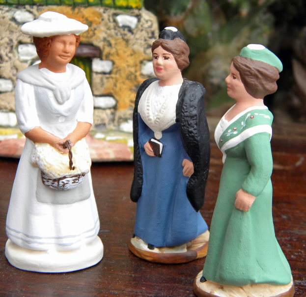 Drei Frauen-Skulpturen-Puppen auf einem Holzbrett mit Spielzeughäusern im Hintergrund.