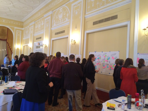 Eine Gruppe von Menschen untersucht Plakate und Diagramme, die an einer Wand in einem Gebäude zu sein scheinen, das wie ein Kunstmuseum aussieht.