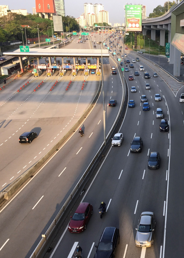 Eine vielbefahrene Autobahn mit zahlreichen Autos, Motorradfahrern, Verkehrskegeln, Verkehrszeichen, Laternen, einer Brücke, Bäumen, Gebäuden und einem klaren blauen Himmel.