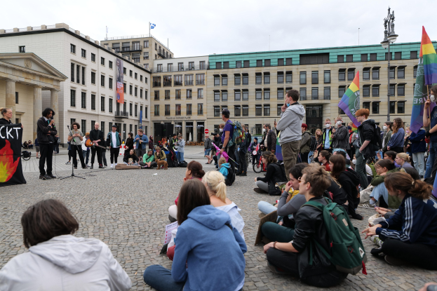 Eine Gruppe von Menschen, die auf dem Boden vor einer Menge sitzen, die Fahnen und Schilder hält, während einer anti-schwulen Demonstration in Berlin, mit einer Statue, Gebäuden und einem bewölktem Himmel im Hintergrund.