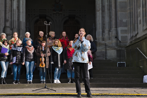 Eine Gruppe von Menschen, einige mit Musikinstrumenten, steht vor einem Gebäude mit Treppen und Geländern, während eine Frau in der Mitte in ein Mikrofon singt, umgeben von Mikrofonständen.