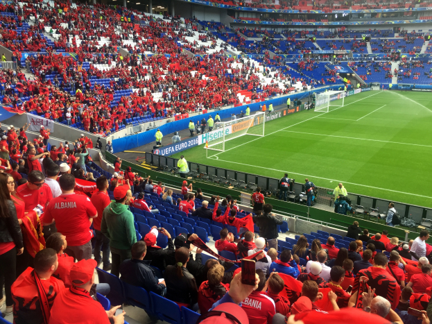 Ein volles Stadion mit Zuschauern, einige sitzen, einige stehen, mit Fahnen, einem Torpfosten und Deckenbeleuchtung, die gespannt auf den Beginn eines Fußballspiels warten.