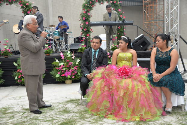 Ein Mann steht und spricht in ein Mikrofon, während andere sitzen und ein Orchester im Hintergrund spielt, auf einer Bühne, die mit Blumen und Pflanzen geschmückt ist.