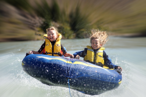 Zwei Kinder in Schwimmwesten sitzen auf einem aufblasbaren Reifen im Wasser, mit einem Seil, das am Reifen befestigt ist, vor einem unscharfen Hintergrund.
