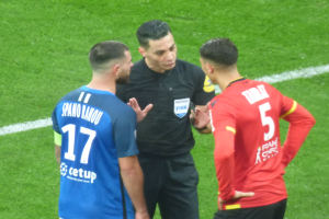 Ein Schiedsrichter in schwarzer Kleidung spricht mit zwei Männern auf einem Fußballfeld, einer in blauen und roten T-Shirts mit Text, beide tragen Brillen.