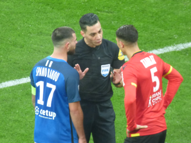 Ein Schiedsrichter in schwarzer Kleidung spricht mit zwei Männern auf einem Fußballfeld, einer in blauen und roten T-Shirts mit Text, beide tragen Brillen.