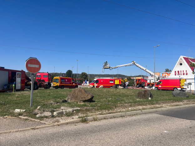 Eine Gruppe von Feuerwehrautos steht auf der Seite einer Straße, umgeben von Gras, Steinen, einem Schild, einem Haus, Polen, einem Kran, Drähten, Bäumen und einem bewölkten Himmel, wahrscheinlich nach einem Feuer an einer Tankstelle.