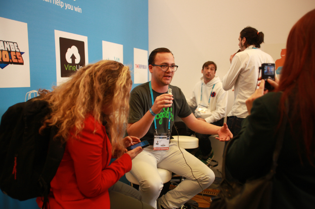 Ein Interviewbild mit Menschen mit Mikrofonen und einer Kamera, einem Banner links und Menschen vorne und in der Mitte, mit einem klaren Hintergrund.