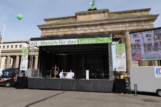 Eine Bühne vor dem Brandenburger Tor in Berlin, Deutschland, mit einem Tisch mit einer Blumenvase, einem Kreuzsymbol und Schildern sowie Lautsprechern und anderem Equipment, mit Fahrzeugen auf der Straße und Gebäuden im Hintergrund.