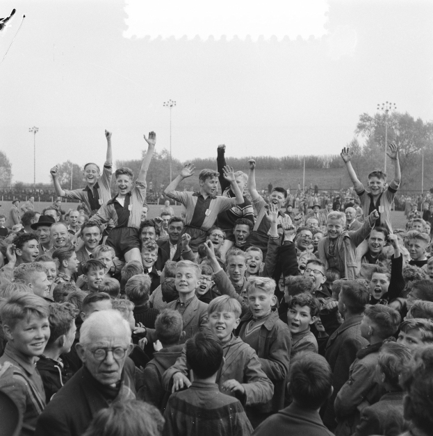 Schwarzes und weißes Foto einer Menge vor einem Stadion, einige mit erhobenen Händen feiernd, mit Laternen, Bäumen und einem klaren Himmel im Hintergrund.