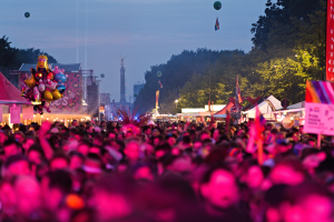 Eine große Menschenmenge auf einem nächtlichen Musikfestival, beleuchtet von farbigen Lichtern und Ballons, mit Zelten, Bannern, Fahnen und anderen Gegenständen, die herumliegen, Bäumen, einem Turm und einem sternenklaren Himmel im Hintergrund.