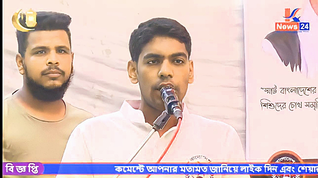 Zwei Männer stehen nebeneinander vor einem Fernseher, einer spricht in ein Mikrofon in einem weißen Hemd, der andere in einem hellgrauen T-Shirt, mit einem Banner und Wasserzeichen im Hintergrund.