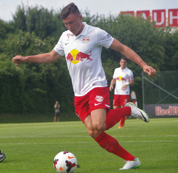 Ein professioneller Fußballspieler schießt einen Ball auf einem Rasenfeld mit Bäumen und einem klaren blauen Himmel im Hintergrund, mit "RB Leipzig" auf einer Tafel dahinter.