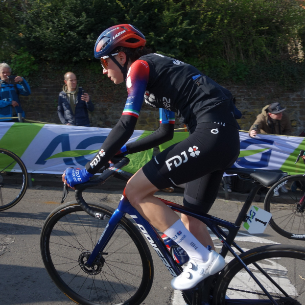 Professionelle Radrennfahrerin Katie Archibald mit Helm, Schutzbrille, Handschuhen und Schuhen auf einem Fahrrad die Straße entlang fahrend, neben einer Menschenmenge, mit einem Banner, einer Wand, Bäumen und einem Gebäude im Hintergrund.