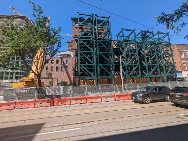 Große Metallbrücke im Bau an einer Baustelle mit parkenden Autos, Zaun, Bäumen, Gebäuden, einem Kran und Himmel im Hintergrund.