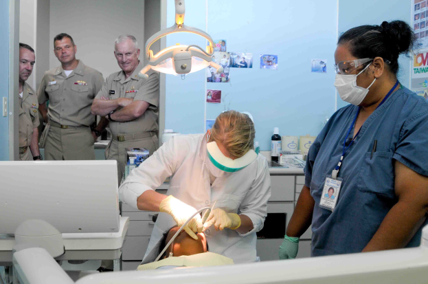 Eine Gruppe von Menschen in einer Zahnarztpraxis, mit einer Person in einem weißen Kleid, die ein Objekt hält, einer weiteren mit Brille, Maske und Ausweis, sowie Plakate an der Wand im Hintergrund.