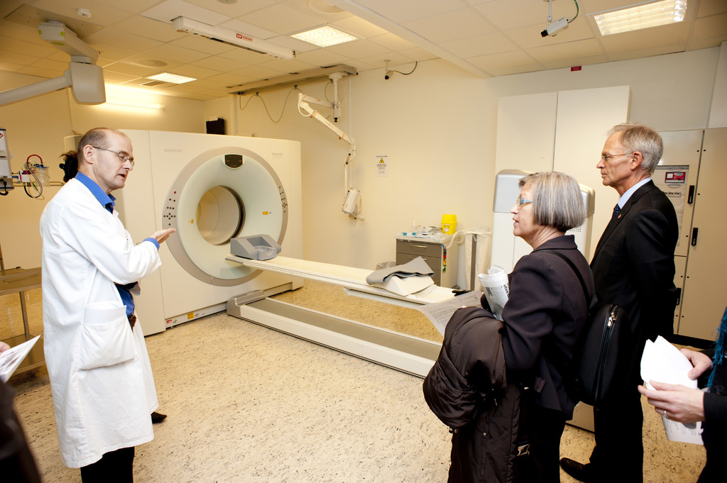 Ein Arzt in weißer Uniform erklärt eine CT-Scan-Maschine zwei Personen in schwarzen Mänteln, die aufmerksam zuhören.