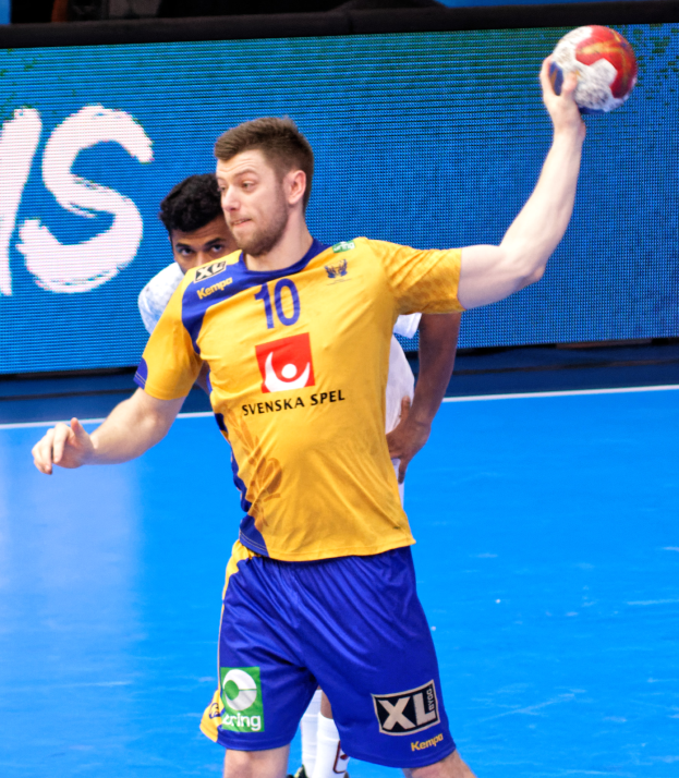 Ein Mann in einem gelben T-Shirt und blauen Shorts spielt Handball auf einem Platz, hält einen Ball und hat im Hintergrund einen Bildschirm mit der Aufschrift "Svenský Spel Handball World Cup 2019".