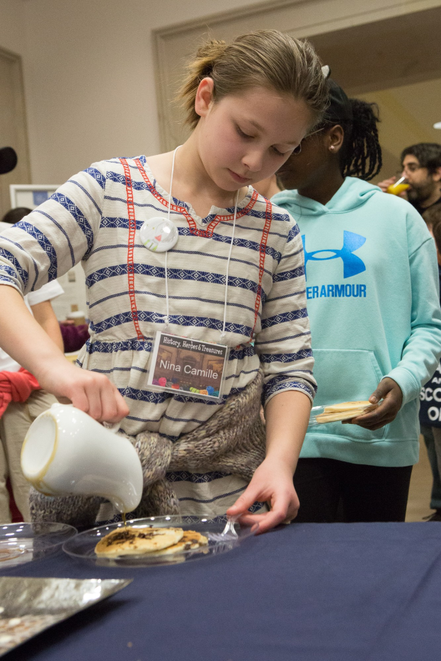 Ein junges Mädchen gießt Milch aus einem Krug auf einen Teller auf einem Tisch, umgeben von stehenden Menschen, mit zusätzlichen Tellern und einem Tablett auf dem Tisch und einer Wand mit einer Tür im Hintergrund.