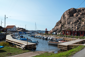 Ein Hafen mit Booten, die an einem felsigen Ufer festgemacht sind, eine Straße mit Gras auf beiden Seiten links und Häuser, Pfosten, Straßenlaternen, Fahrzeuge, Felsen und einen klaren blauen Himmel im Hintergrund.