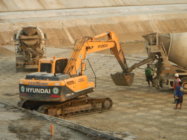 Eine Baustelle mit einem Bagger und einem Betonmischer im Vordergrund und einigen Menschen in der Nähe, mit einer Wand im Hintergrund.