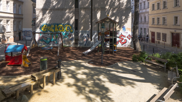 Spielplatz mit Graffiti, umgeben von Gebäuden, Bäumen, Bänken und einem Mülleimer, mit Menschen und Fahrzeugen auf der Straße und sichtbarem Himmel im Hintergrund.