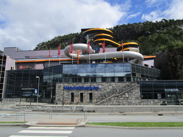 Das Handelsbanken Museum in Norwegen, ein großes Gebäude mit zahlreichen Fenstern, umgeben von einer Straße, Gras, Geländern, Pfählen, Laternen, Schildern, Treppen und anderen Gegenständen, mit Bäumen und einem bewölkten Himmel im Hintergrund.