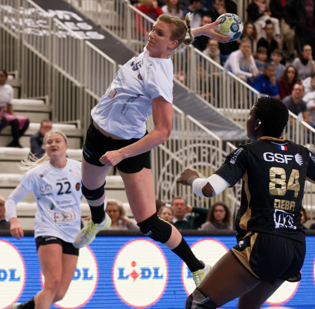 Gruppe von Frauen, die auf einem Platz Handball spielen, mit einem Ball in der Luft, Zuschauern auf Treppen und einer "Handball-Weltmeisterschaft 2019 - Tag 2"-Tafel im Hintergrund.