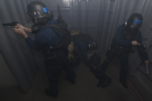 Polizisten in Einsatzausrüstung mit einem, der eine Waffe hölt, neben einem Metallbehälter und einer Leiter in einem begrenzten Raum.