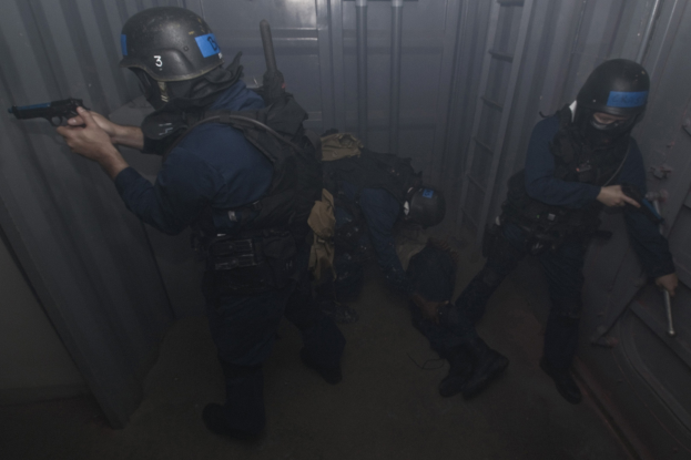 Polizisten in Einsatzausrüstung mit einem, der eine Waffe hölt, neben einem Metallbehälter und einer Leiter in einem begrenzten Raum.