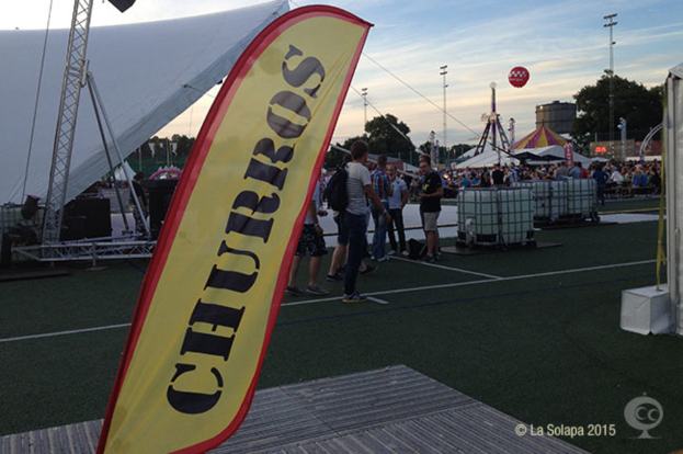 Eine KarnevalsSzene mit Zelten, einem zentralen Mast, Menschen, Bäumen, Strommasten, Straßenbahnen auf der Straße und einem eisernen Stand auf der linken Seite unter einem Himmel-Hintergrund.