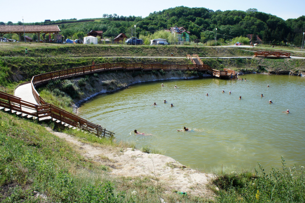 Gruppe von Menschen, die in einem Gewässer schwimmen, mit einer Brücke, Treppen, Schuppen, Fahrzeugen, Pfählen und üppiger grüner Umgebung unter einem klaren blauen Himmel.