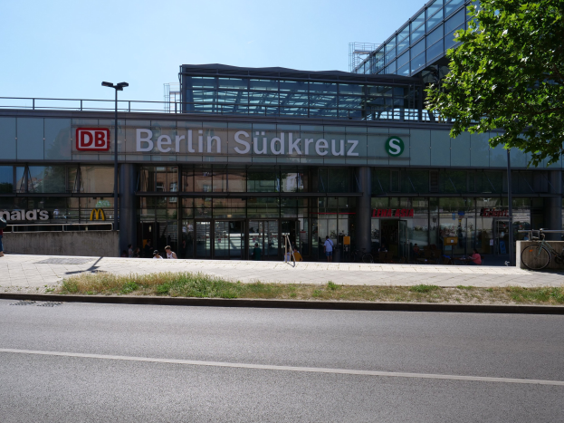 Großes Glasgebäude des Berliner Südkreuz-Bahnhofs mit Text an seiner Fassade, umgeben von Straßenlaternen, Lichtern, Fahrzeugen, Fußgängern, Fahrrädern, Bäumen und einem klaren blauen Himmel.
